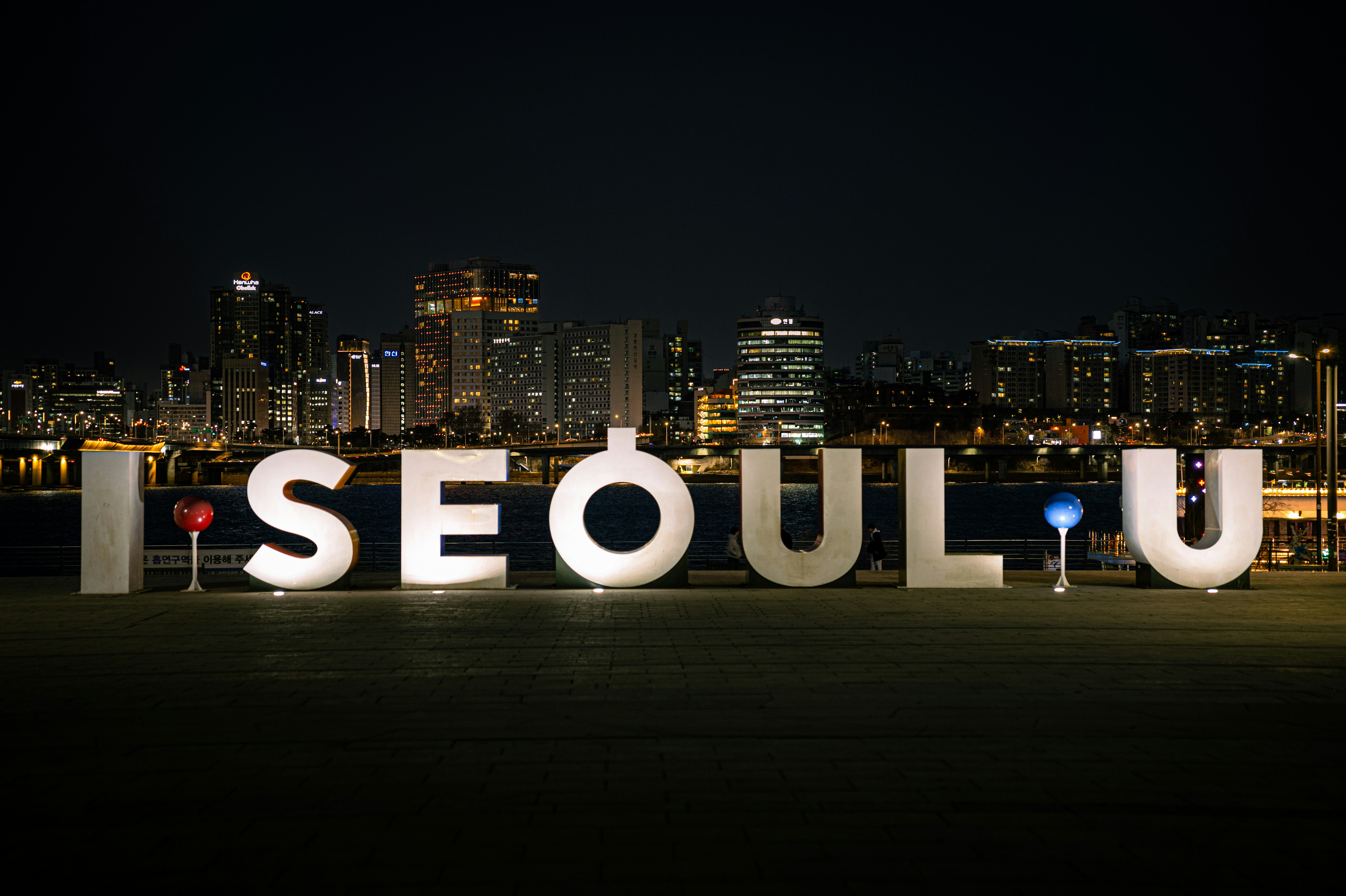 发光的“I SEOUL U”标志在夜晚的汉江边熠熠生辉，首尔城市灯光在背景中闪烁。