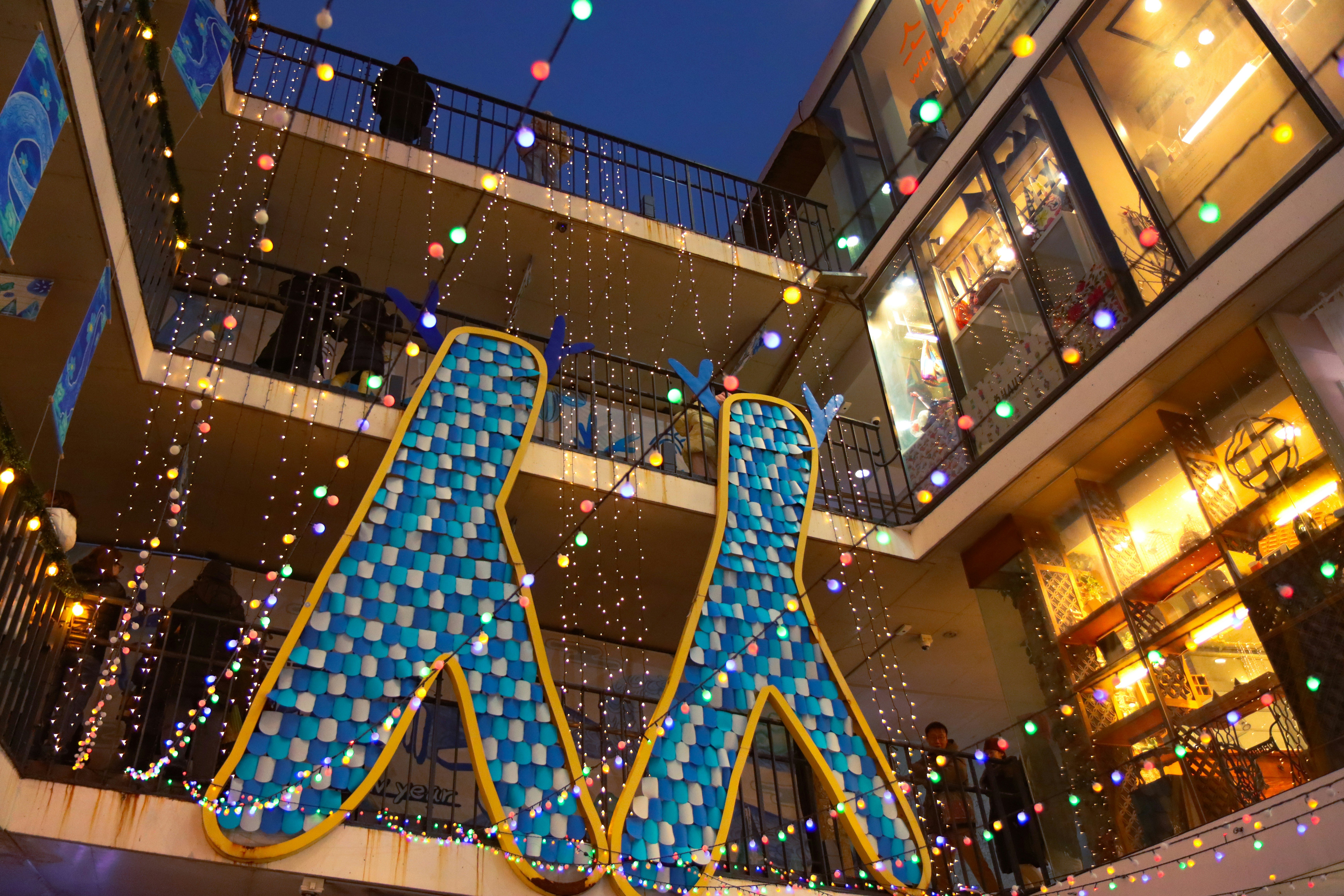 Một khu phức hợp mua sắm đầy màu sắc được thắp sáng bằng đèn dây lễ hội và các vật trang trí hình đôi chân màu xanh lớn treo giữa các ban công vào lúc hoàng hôn.